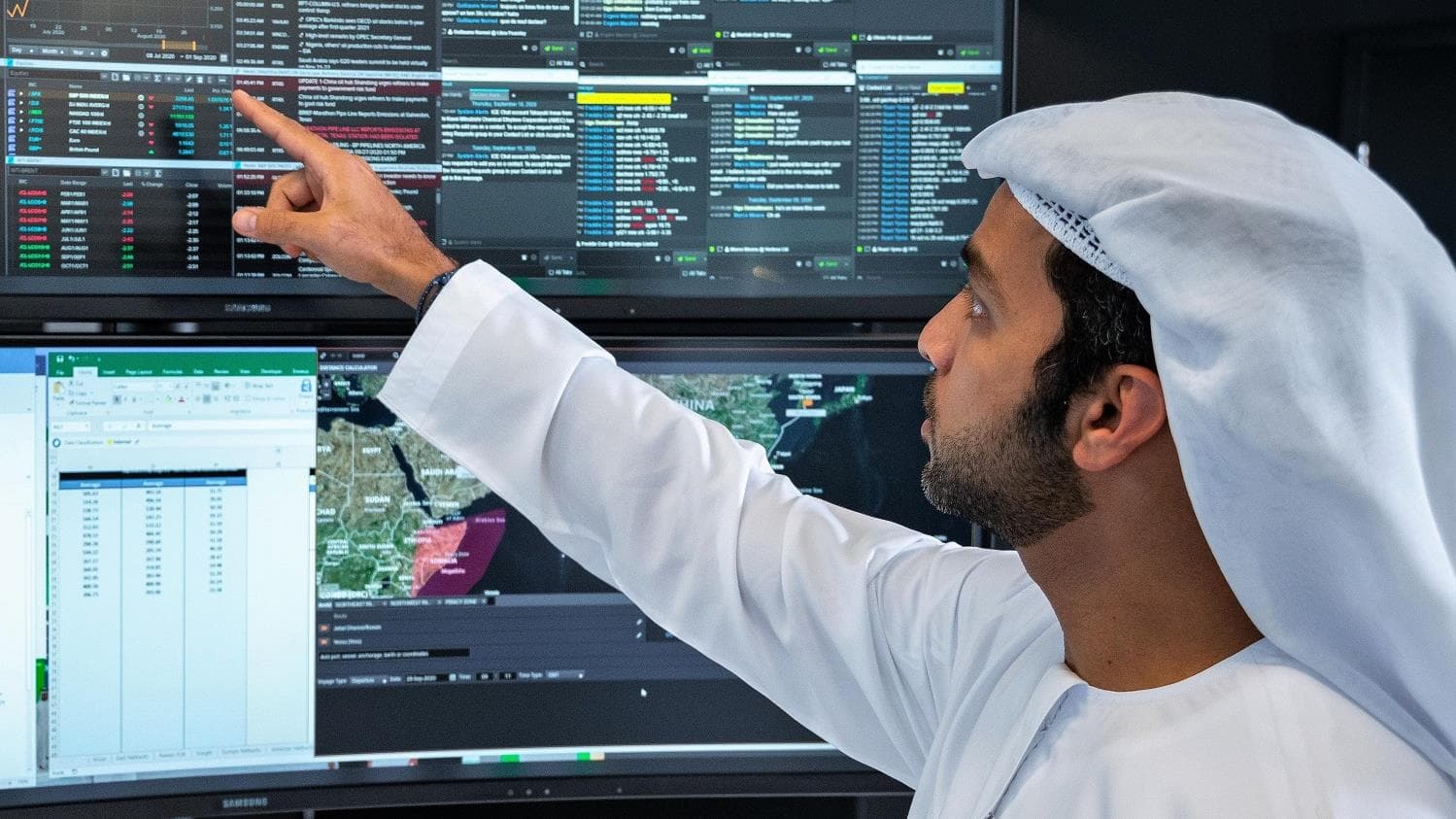 An Emirati trader in traditional dress points at a wall of financial market screens showing prices, charts, and a map of the region