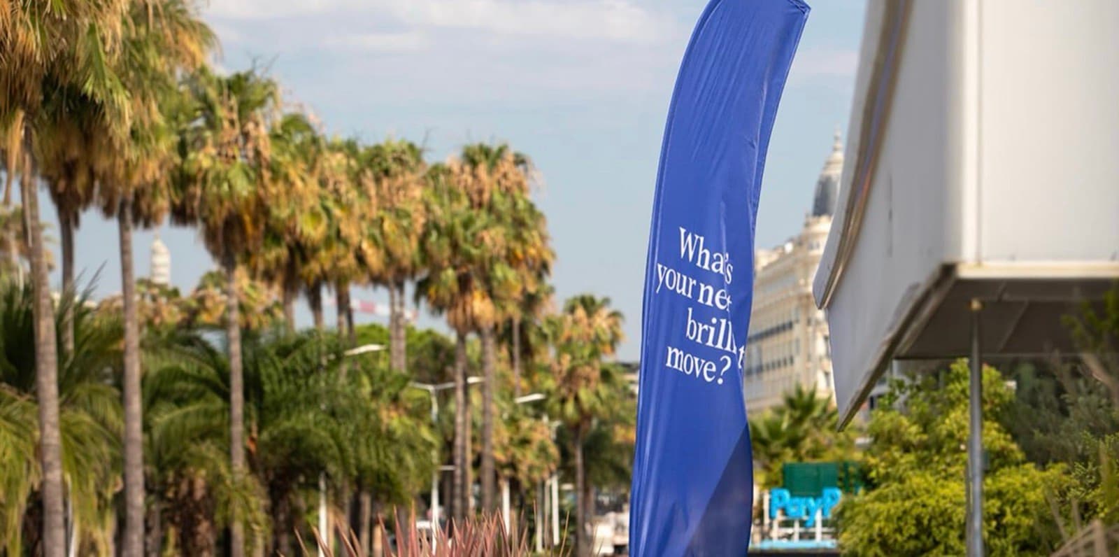 A tall blue Cannes Lions banner on the Croisette reading What's your next brilliant move, with palm trees and a PayPal activation behind it