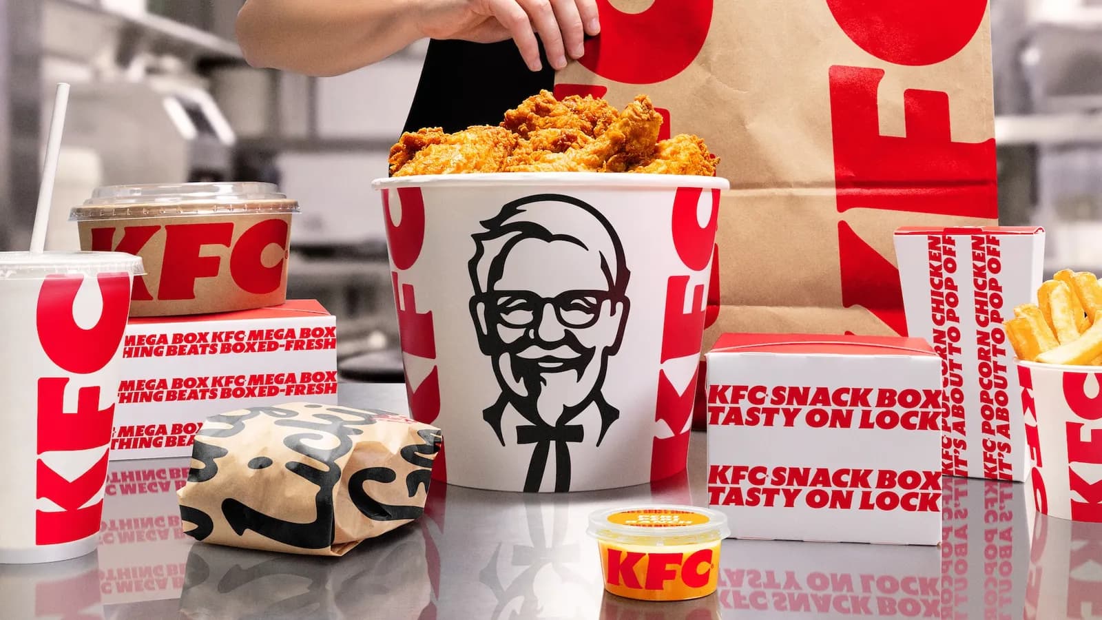 A KFC bucket of fried chicken with the Colonel logo, surrounded by refreshed KFC packaging