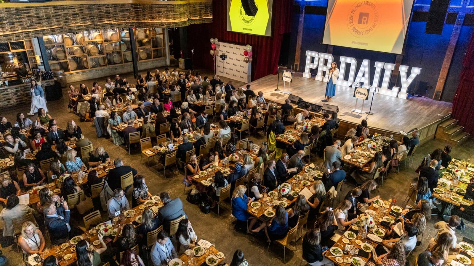 An aerial view of a packed awards luncheon at City Winery, with diners at long tables facing a stage lit by giant marquee letters spelling PR Daily