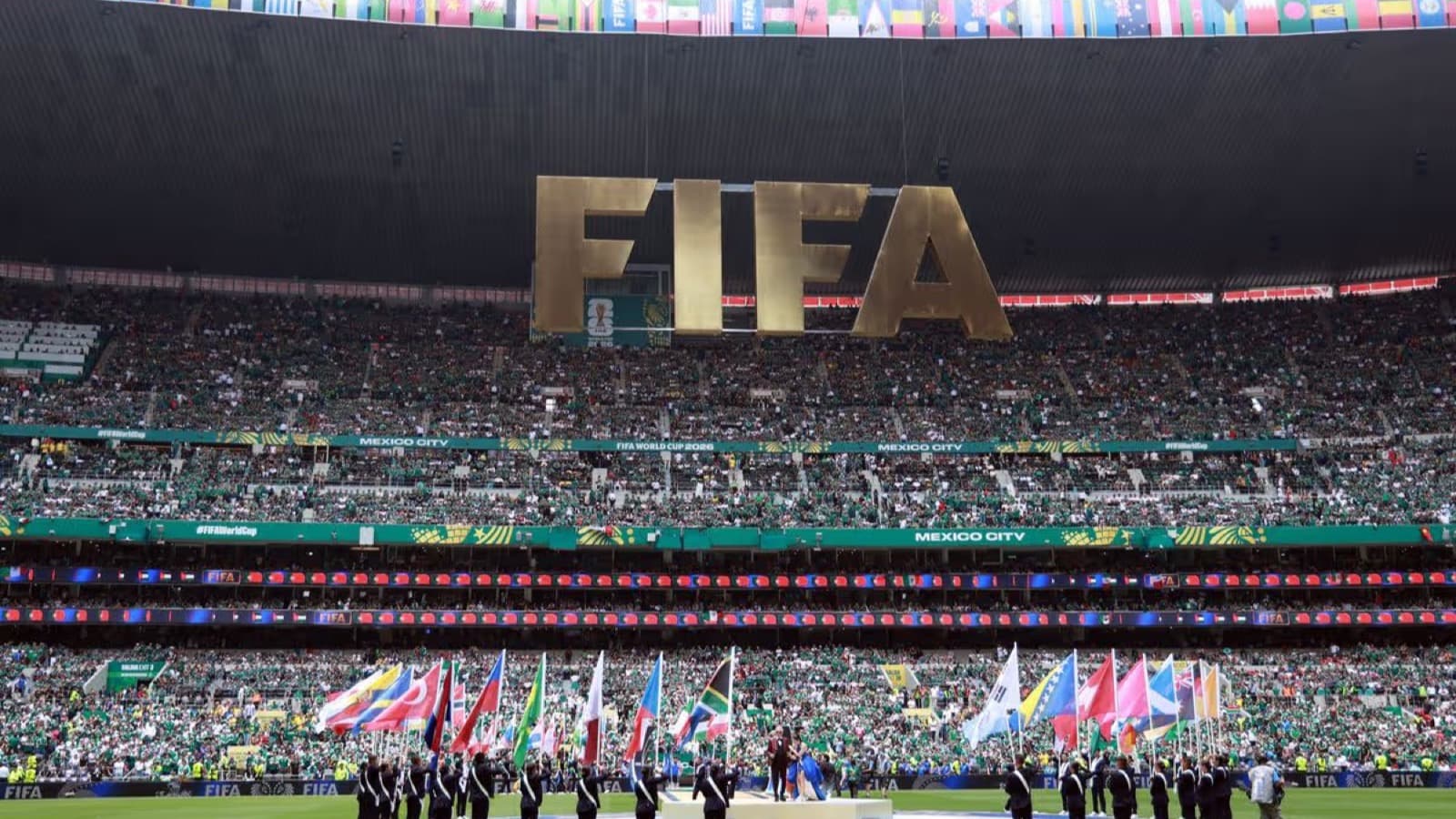 A packed stadium during the 2026 World Cup with a giant FIFA sign and national flags carried across the pitch in Mexico City