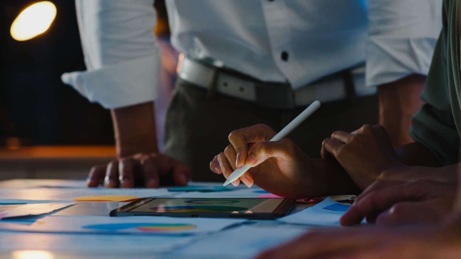 A team of colleagues gathered around a table reviewing charts on a tablet with an Apple Pencil during a brainstorm