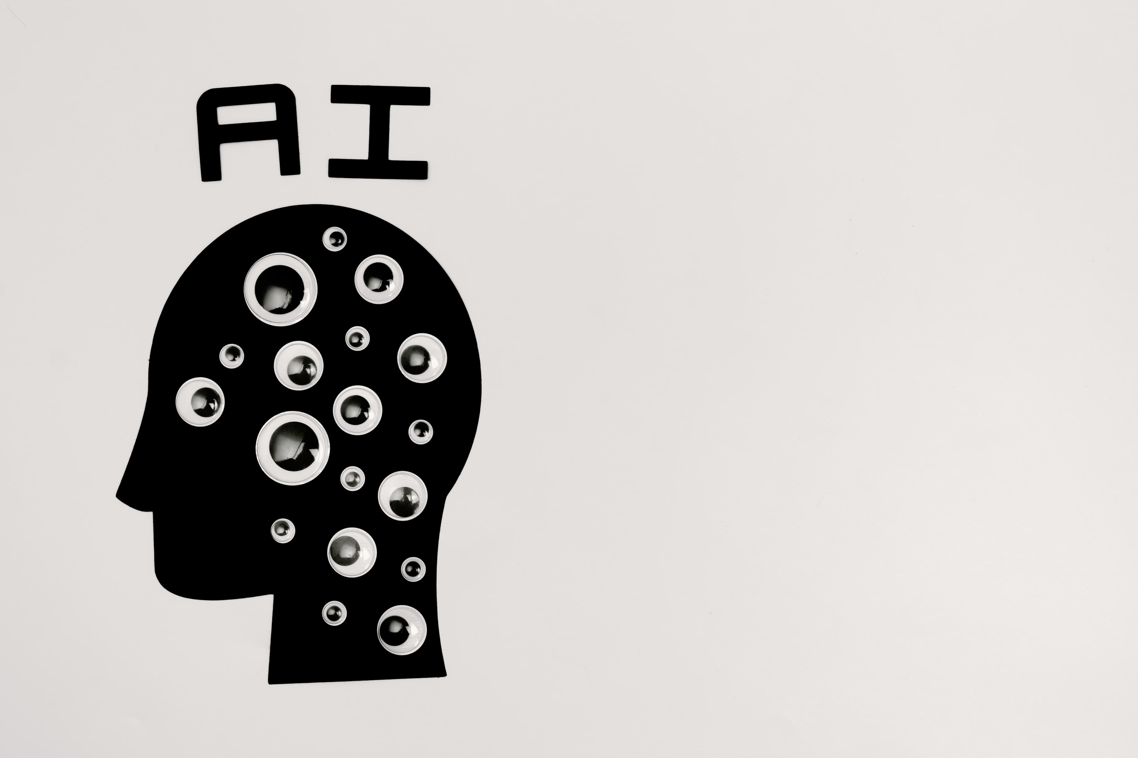 A black paper cutout of a human head labeled AI filled with small round eyes on a white background