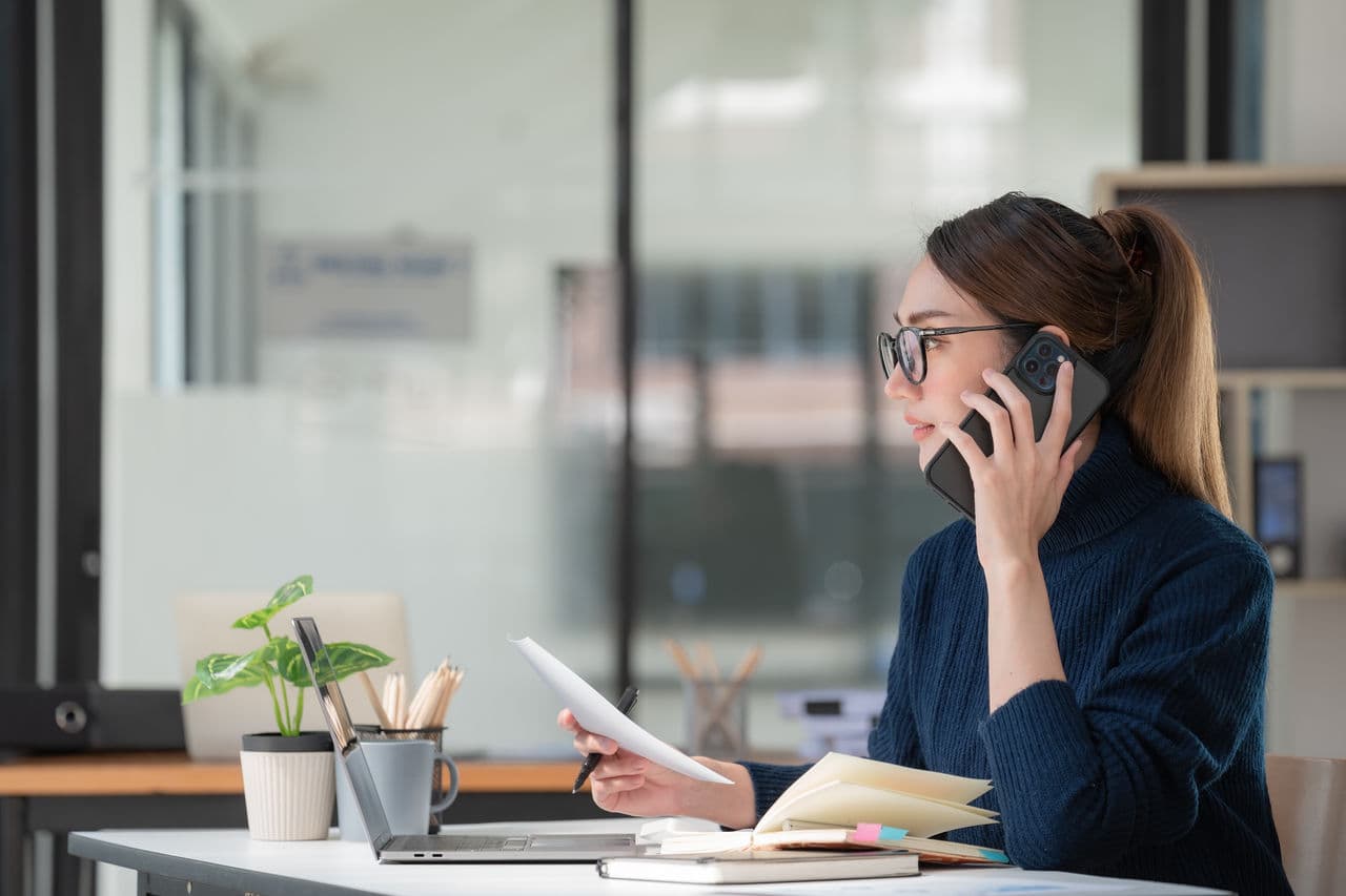 A marketer at a desk on the phone reviewing a document beside a laptop