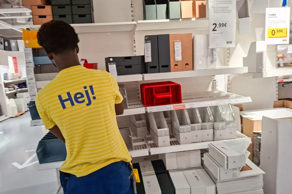 An IKEA employee in a yellow Hej shirt restocking shelves of storage boxes in a store aisle