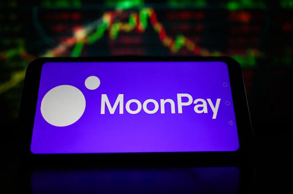 A smartphone displaying the MoonPay logo against a blurred background of stock market charts