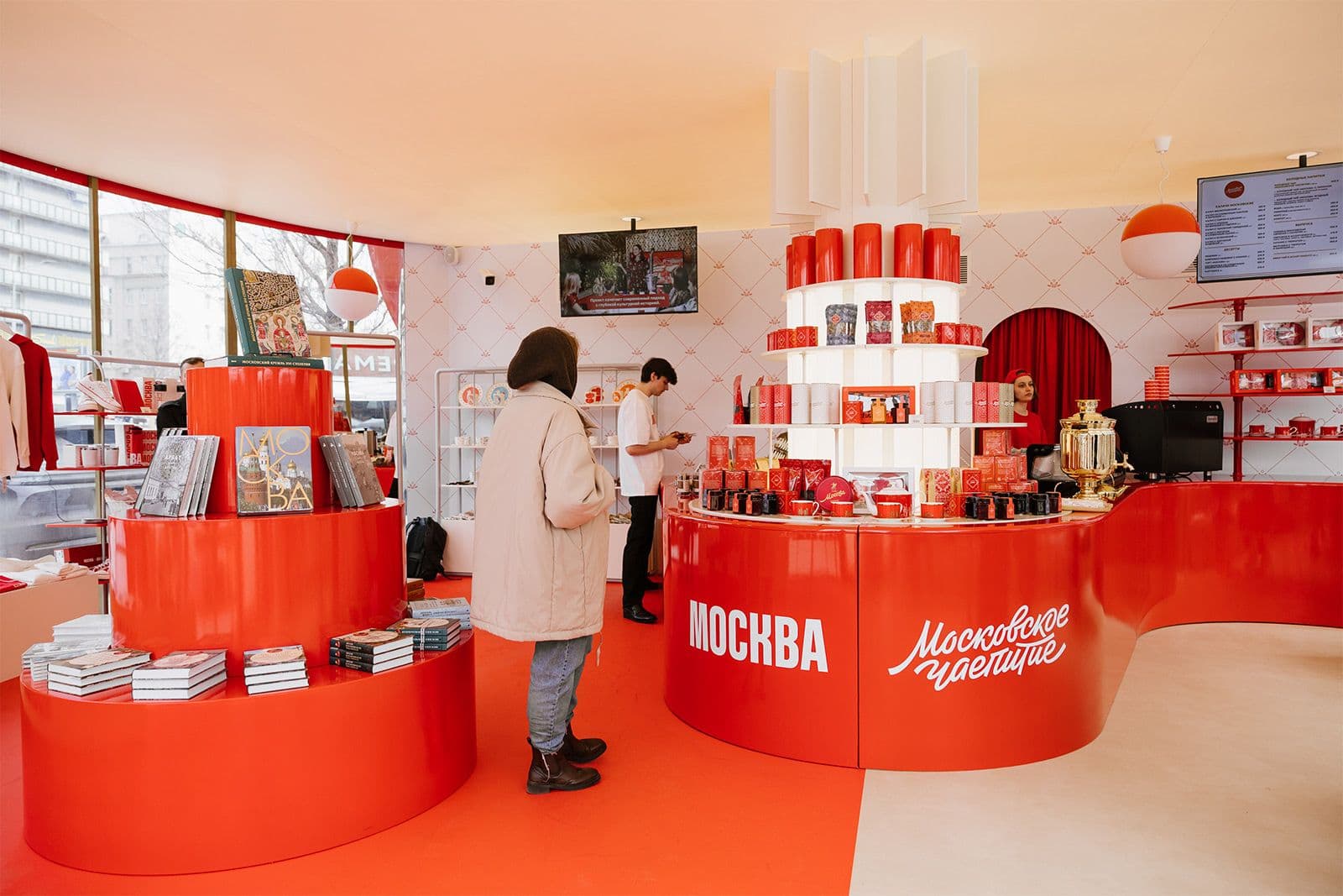 A bright red and white Moscow branded retail pop up displaying Russian books, tea and gift products with a customer browsing