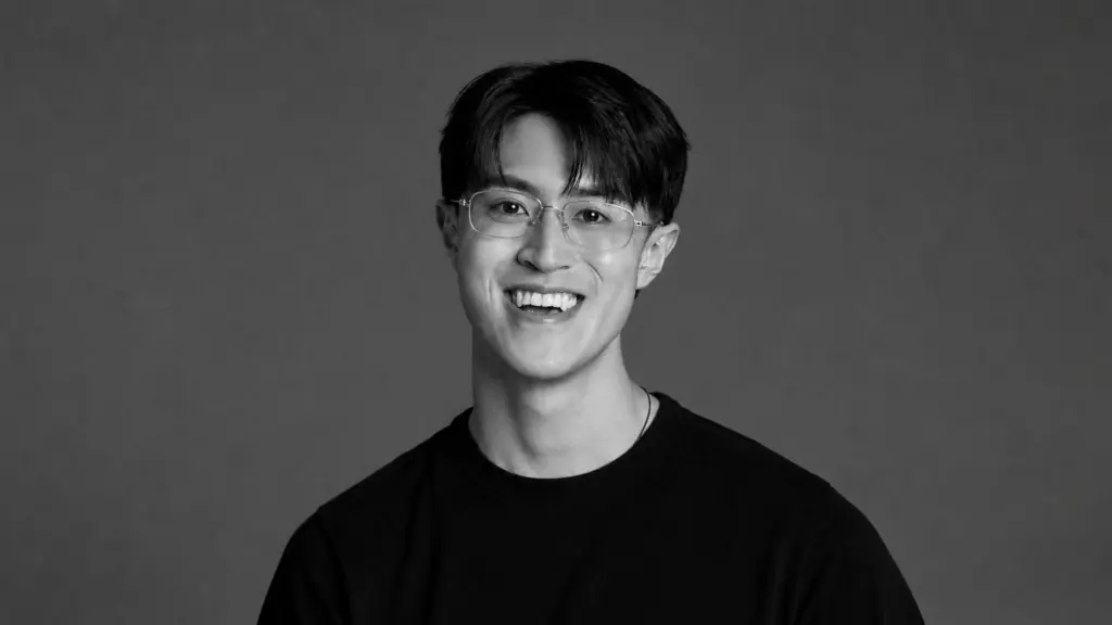 A black and white portrait of a smiling man in glasses and a black t-shirt